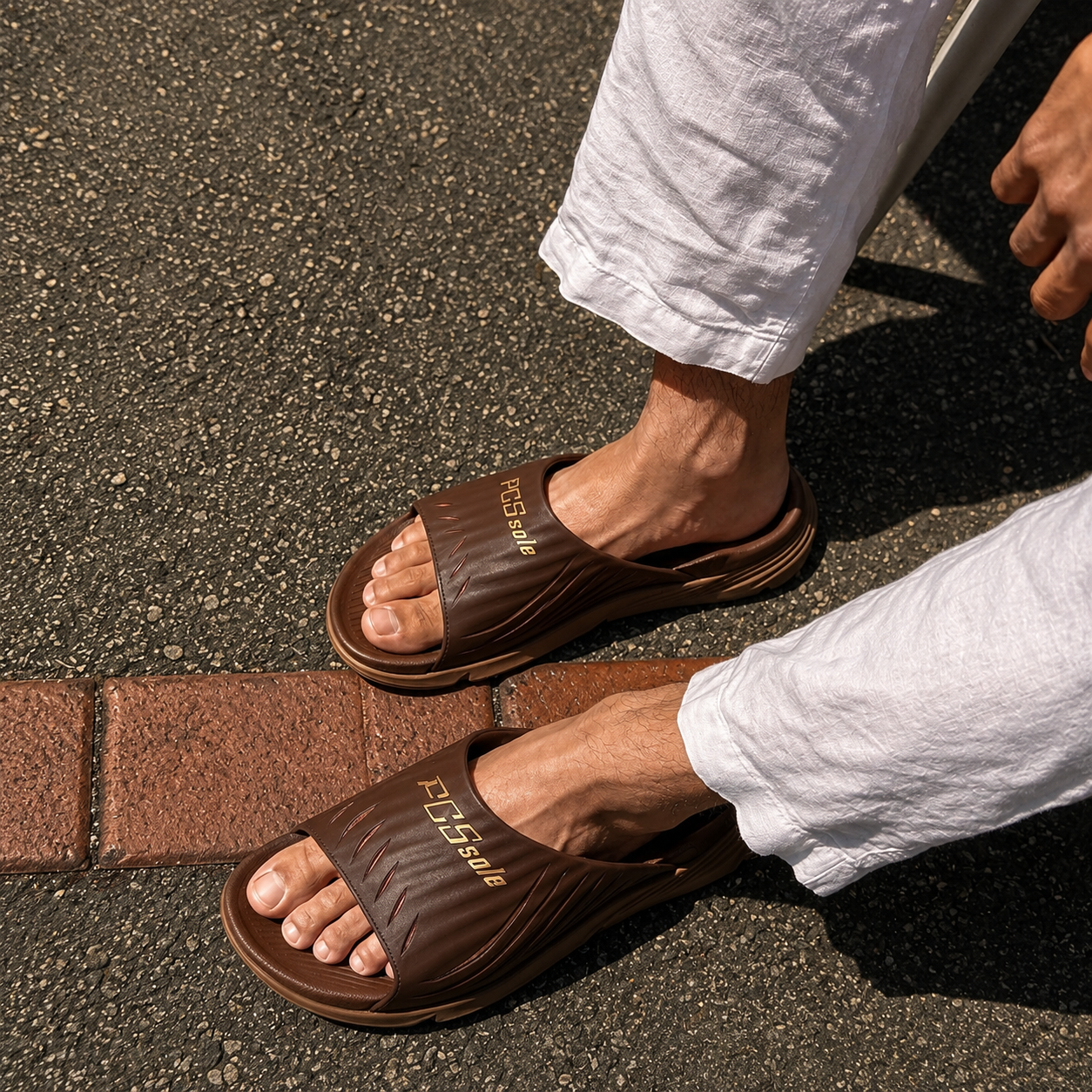 Brown slide sandals worn on a person's feet with white pants on a pavement background