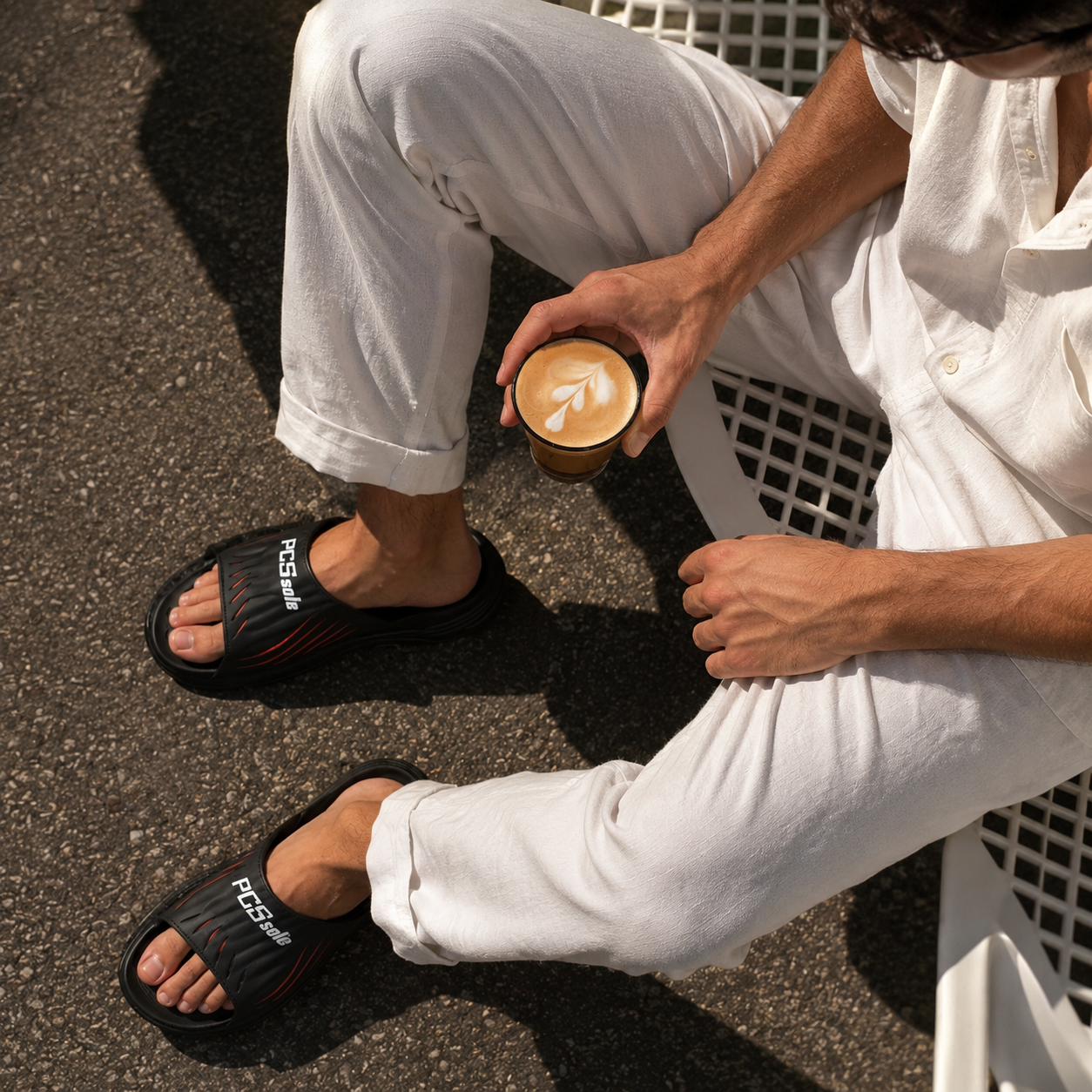 Person wearing black pcssole slides with white text, sitting on a bench holding a coffee cup.