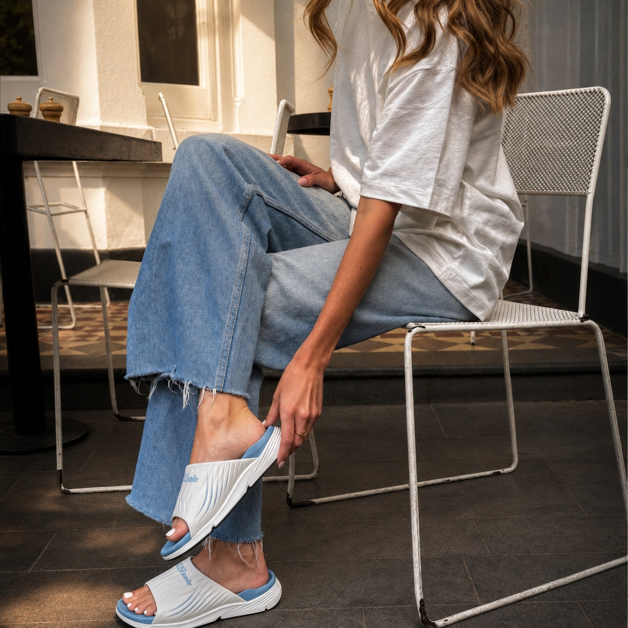 A woman sitting on a white metal chair in a cafe setting, wearing white and blue slide sandals with a white t-shirt and light blue denim jeans.