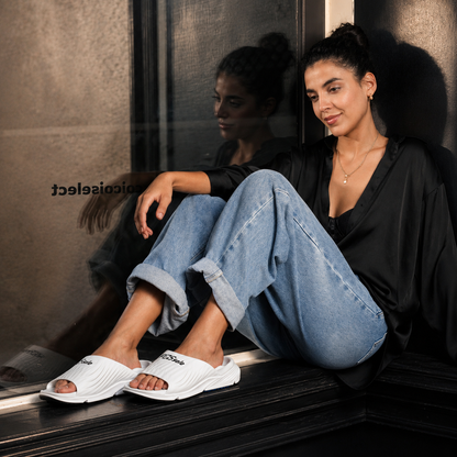 Woman sitting on a ledge wearing white sandals, black top, and blue jeans with a blurred background.