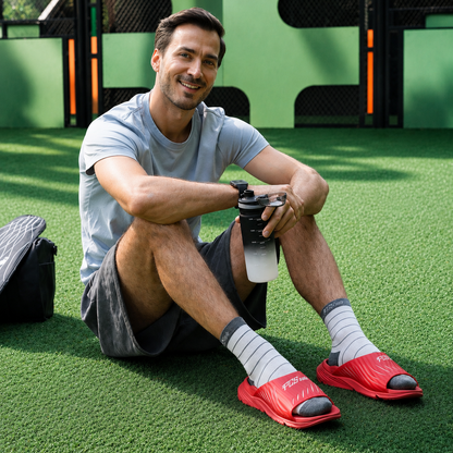 Man sitting on grass wearing red sandals with red soles, holding a water cup.