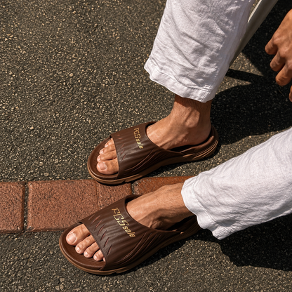 Brown slide sandals worn on a person's feet with white pants on a pavement background