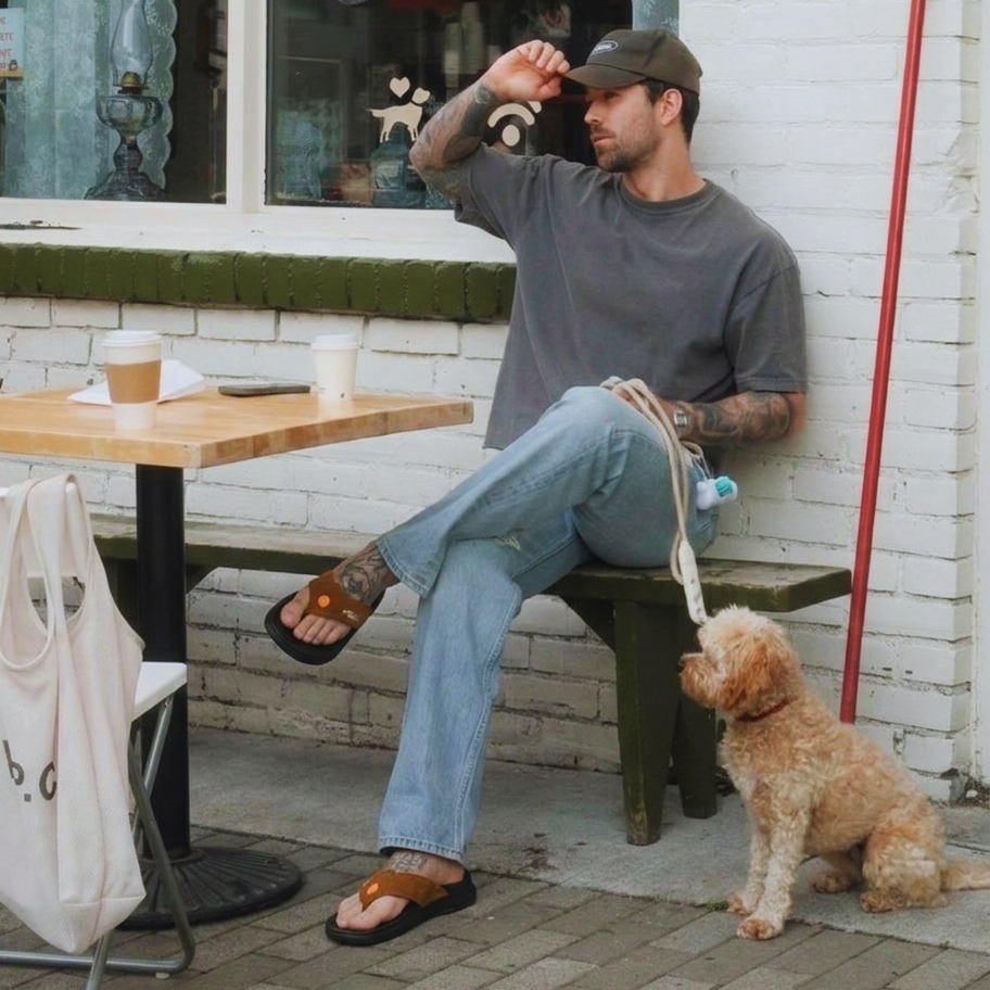 A man wearing a hat and PCSsole Mocha Suede flip-flops sits at an outdoor cafe